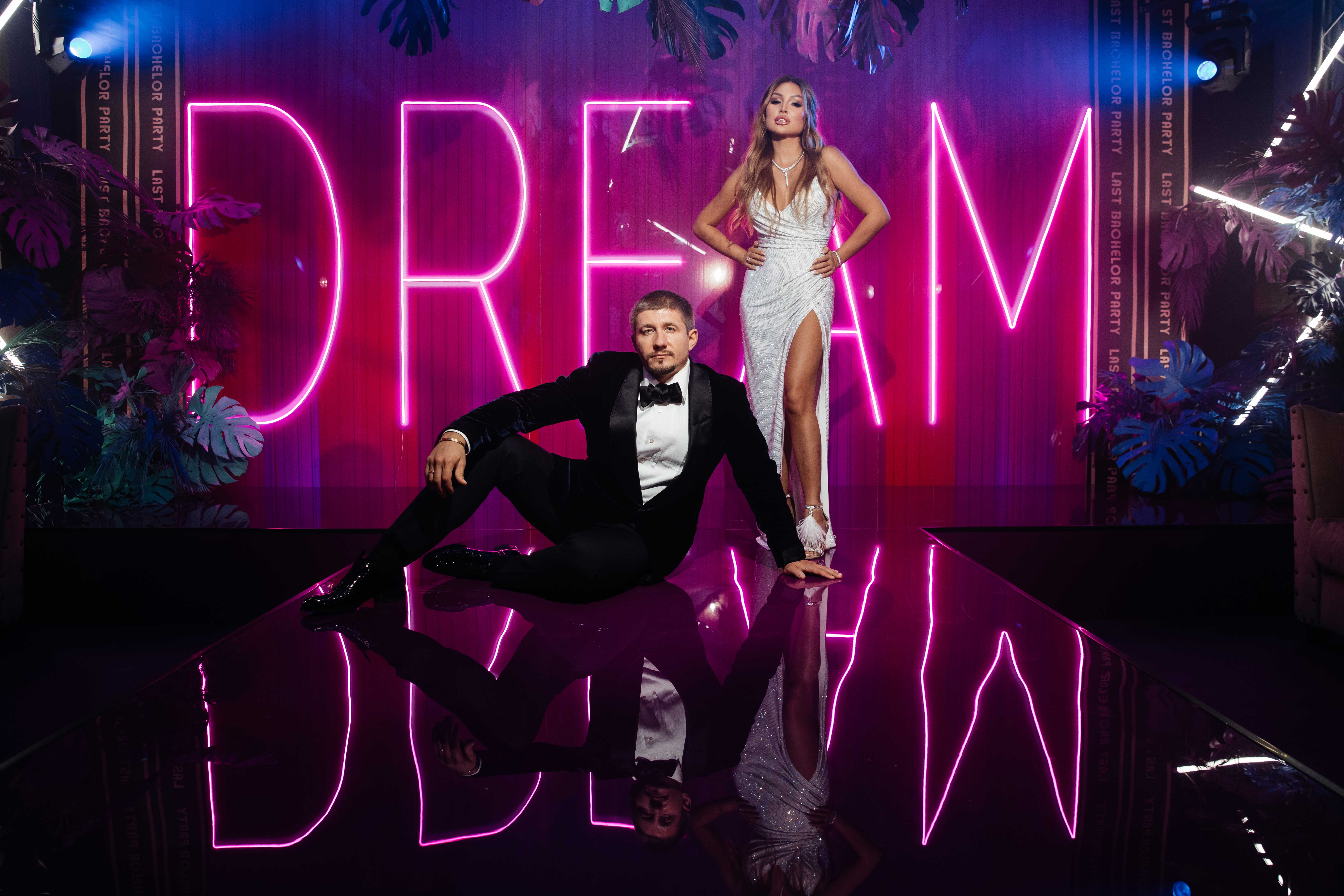 Молодожены на фоне неоновой надписи 'DREAM', мужчина в черном смокинге сидит на полу, женщина в белом вечернем платье стоит рядом