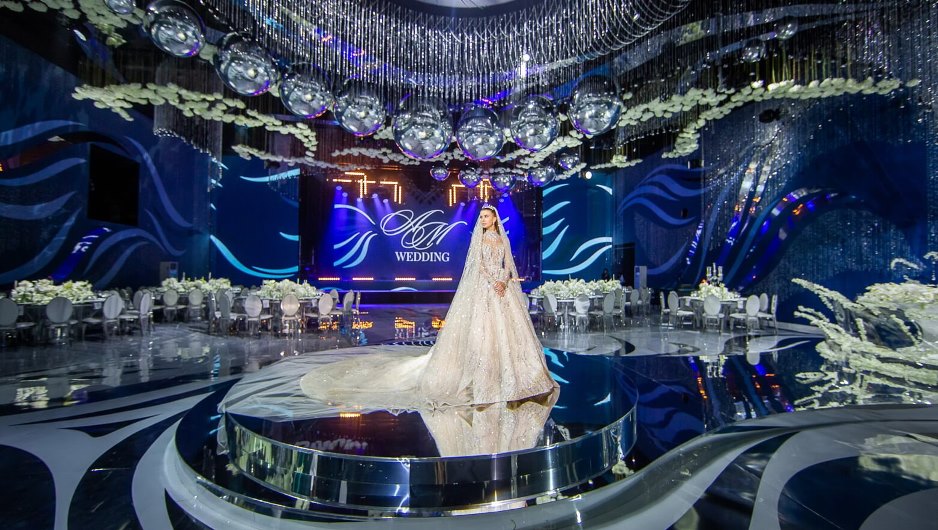 Роскошная свадебная церемония с невестой в великолепном белом платье, стоящей на подиуме в окружении зеркальных шаров и цветочных композиций. Интерьер оформлен в синих тонах с элегантным освещением и